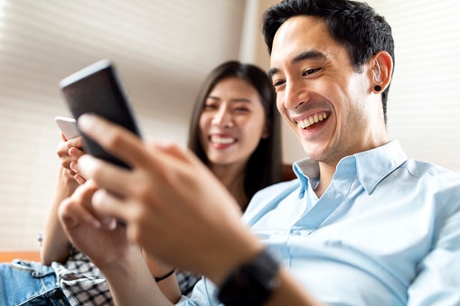 Man and woman on a couch looking at their cell phones and smiling
